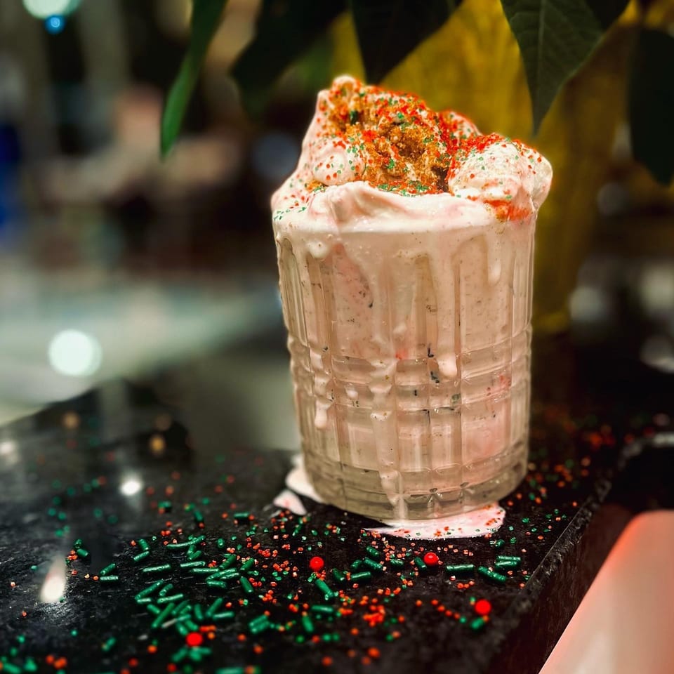 Pink milkshake topped with whipped cream, colorful sprinkles, and crushed cookie pieces in a textured glass on a decorated counter.