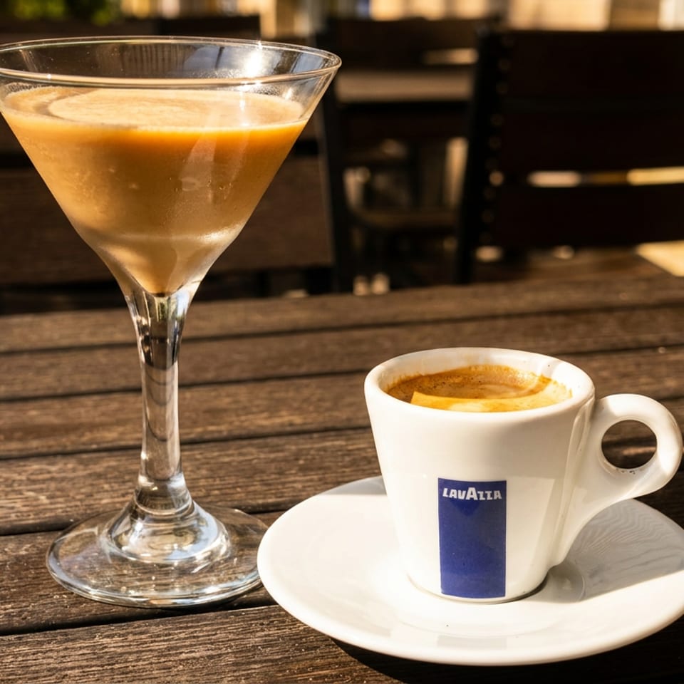 A martini glass and a Lavazza espresso cup on a wooden outdoor table in sunlight.