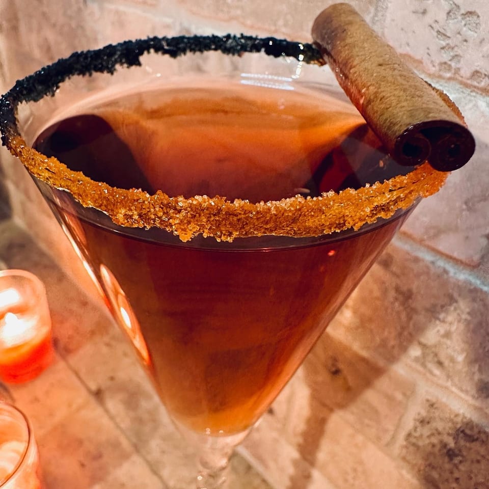 Amber cocktail in a coupe glass with a black-and-gold sugar rim, orange peel garnish, and cinnamon stick.