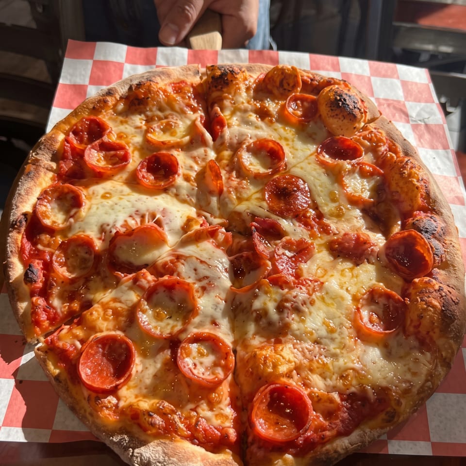 Large pepperoni pizza with melted cheese and charred crust served on red-and-white checkered paper.