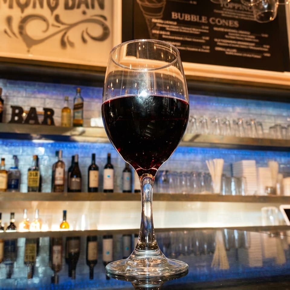 A glass of red wine sits on a reflective bar counter with liquor bottles and glasses in the background.