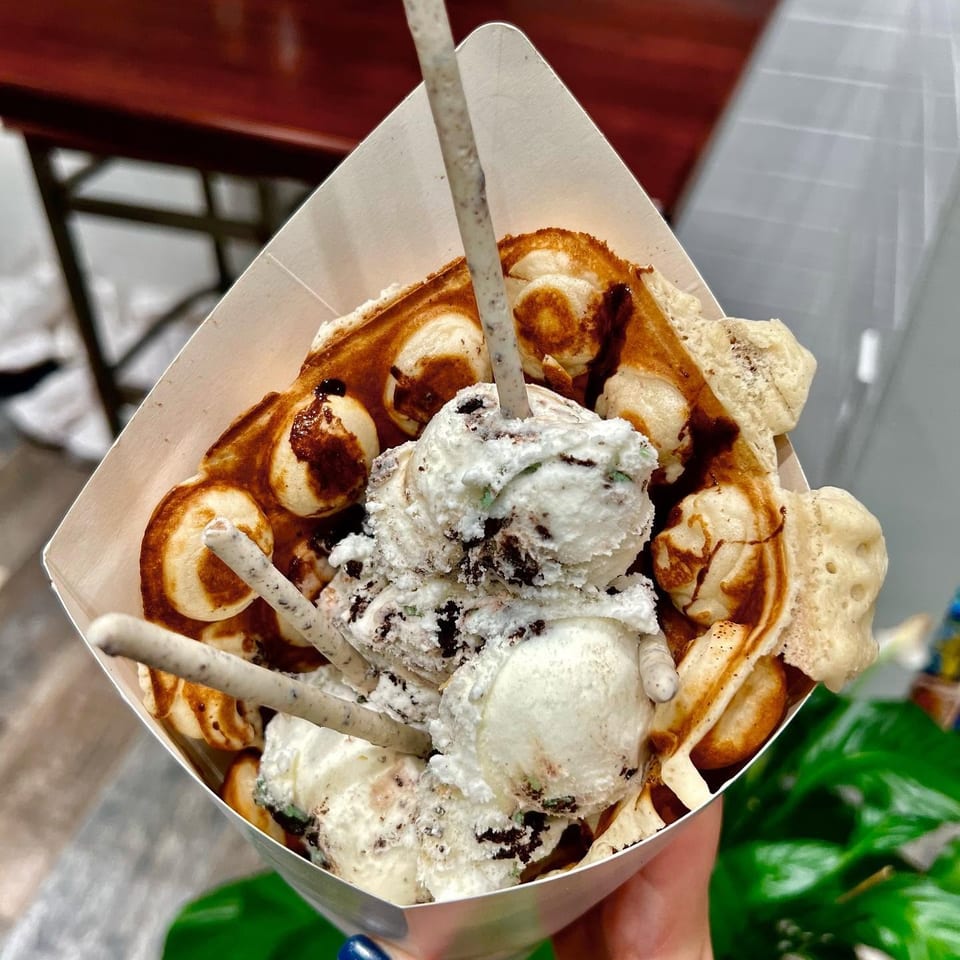 Bubble waffle topped with scoops of cookies-and-cream ice cream, chocolate drizzle, and wafer sticks in a paper cone.