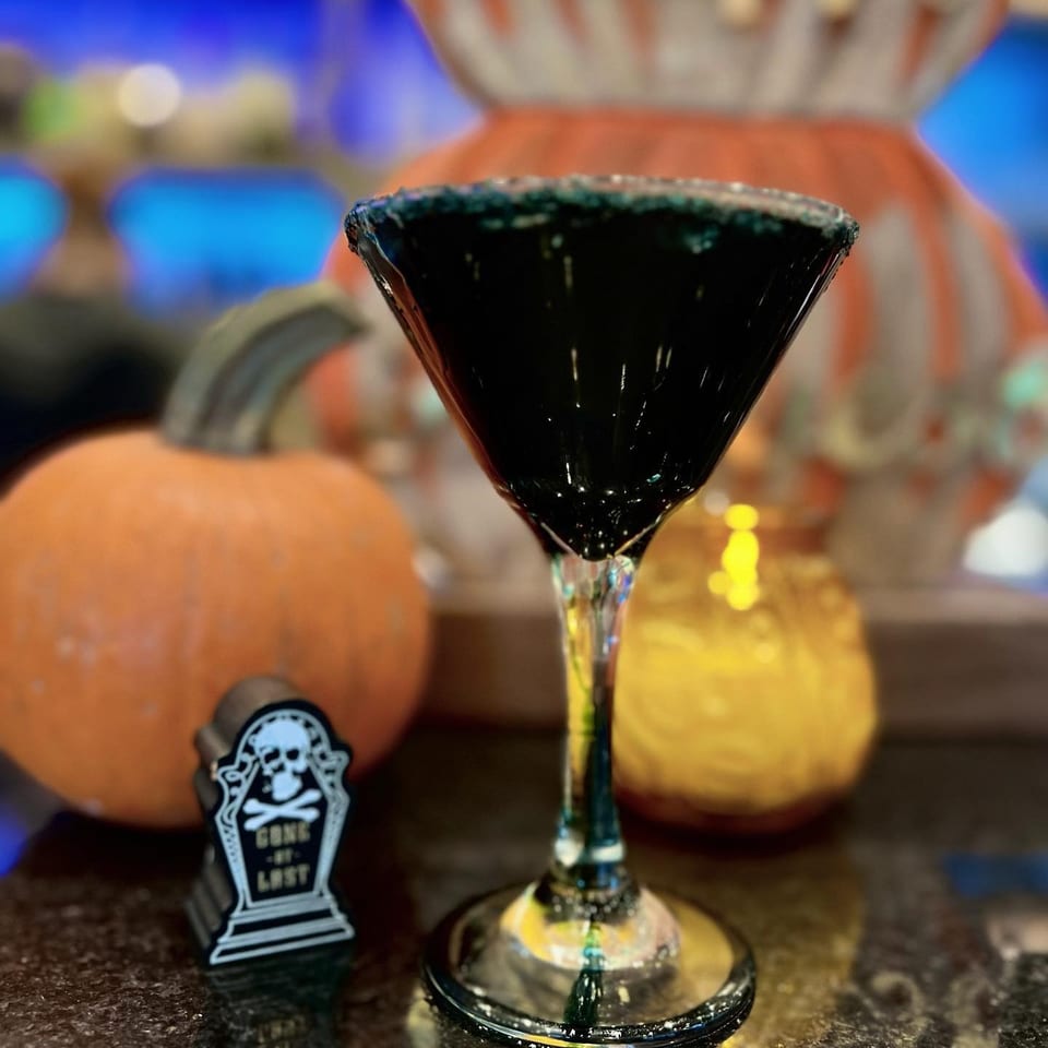 Black cocktail in a martini glass sits on a reflective bar beside pumpkins and a tombstone decoration.