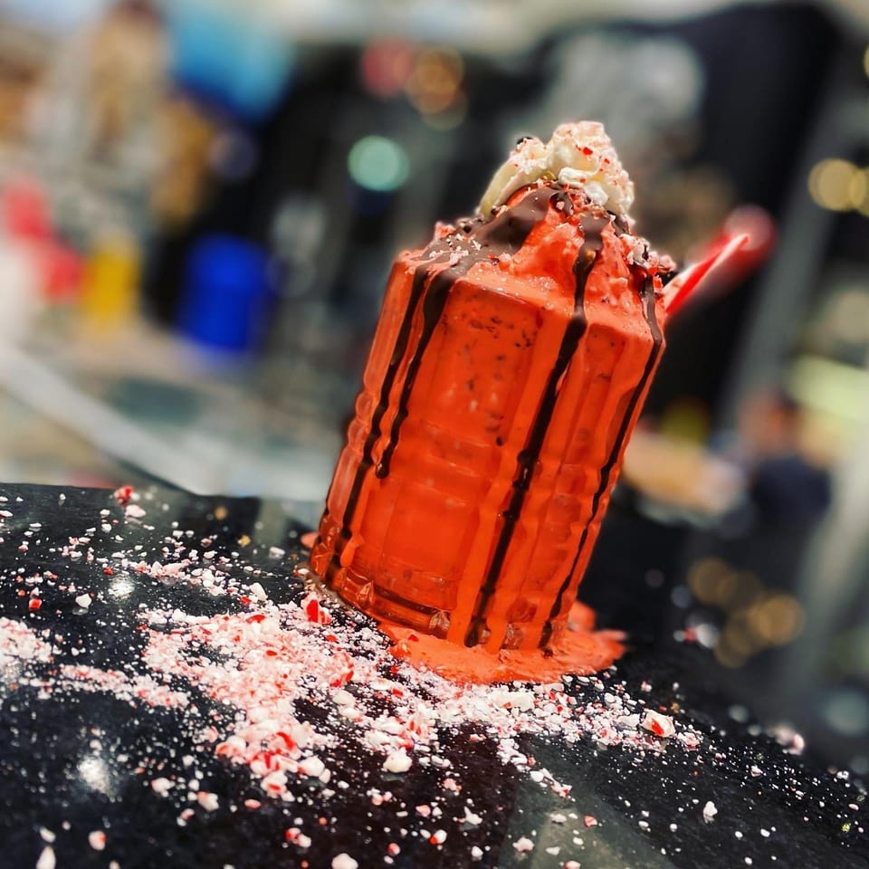 Red dessert drink topped with whipped cream, chocolate drizzle, and crushed peppermint on a black counter.